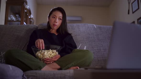 Young Asian woman sitting on sofa and eating popcorn