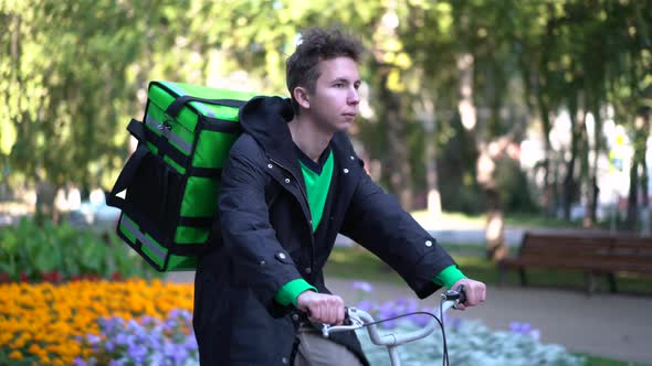 Delivery Man with Green Bagpack Rides a Bicycle Through the City with Food Delivery