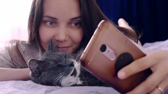 A Young Woman Lies with Her Gray Cat on the Bed Looks at the Smartphone Screen