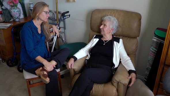 Mature woman and elderly woman talk in a relaxed atmosphere with walker and flowers in the corner.