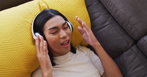 Gender fluid male wearing headphones enjoying listening to music while lying on the couch at home