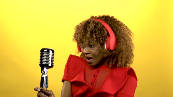 African American Girl in Red Headphones Singing in Retro Microphone. Yellow Background