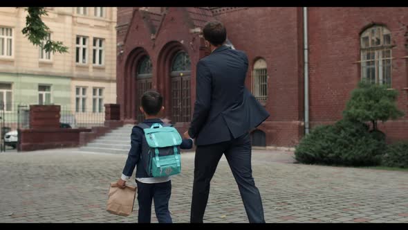 Backside View of Little Kid with Backpack Holding Hand in Hand His Father in Suit. Loving Dad
