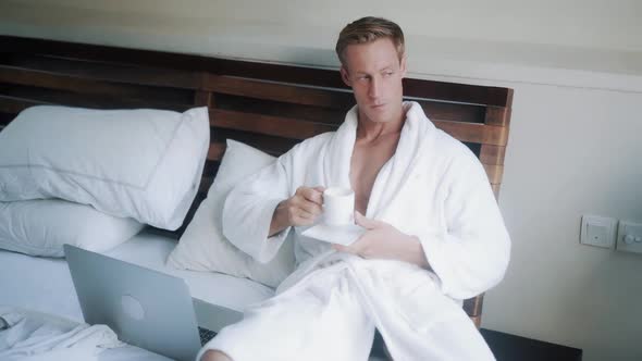 Young Man in Bathrobe Drinks Coffee in Bed Near Laptop