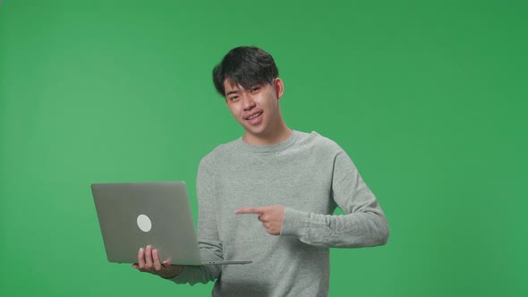 A Smiling Asian Man Pointing To Computer While Standing In Front Of Green Screen Background