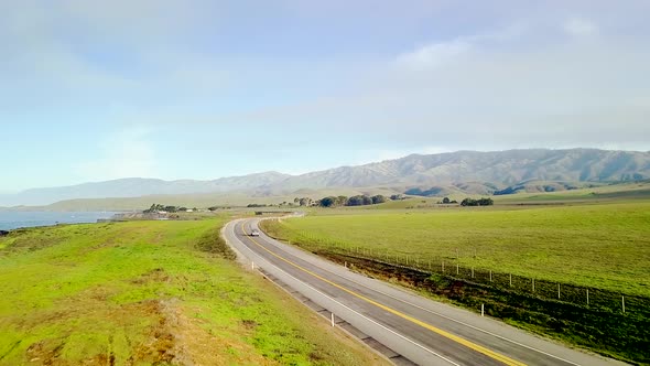 Aerial wide view of winding California coast highway and rolling green hills following fast cars ris