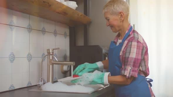 Senior Woman Wearing Apron and Gloves Washing Dishes in Her Kitchen.