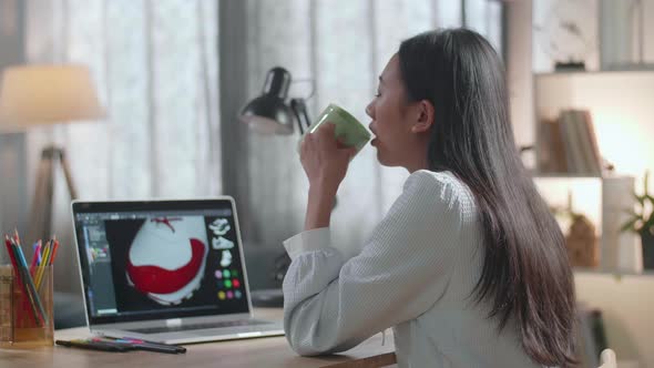 Asian Female Footwear Designer Drinking Coffee While Designing Shoe On A Laptop At Home