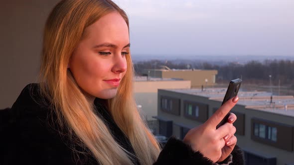 A Young Beautiful Woman Stands on a Balcony and Works on a Smartphone - Closeup