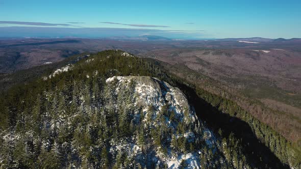 Fast aerial orbit left around the rocky outcrop at the top of a snow dusted mountain in Maine