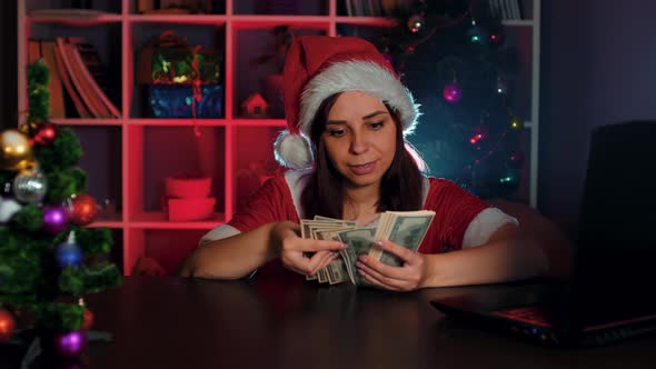 A Young Woman in a Santa Claus Costume Counts Money While Sitting in a Chair in Front of a Computer