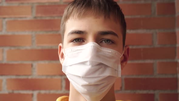 young man in a yellow sweatshirt in a medical facial mask near a brick wall.