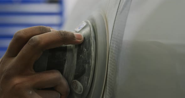 African American male car mechanic wearing a face mask and polishing a side of a car with a grinder