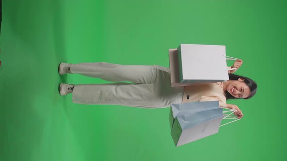 Full View Of Asian Woman Holding Shopping Bags Up Before Posing And Smiling On Green Screen