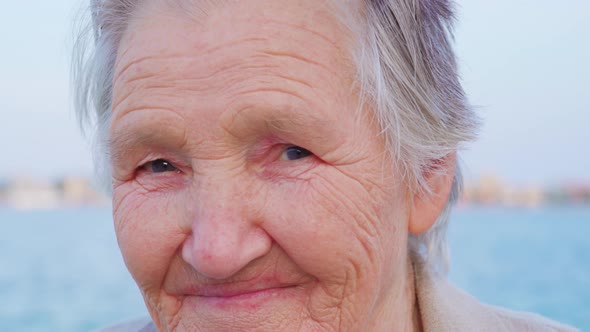 Greyhaired Lady Traveler Stands at Sea Looking Into Camera