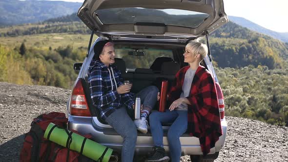 Two Happy Hipster Girls sit in the Trunk of a Car Drinking Hot Coffee or Tea from a Thermos, Talking