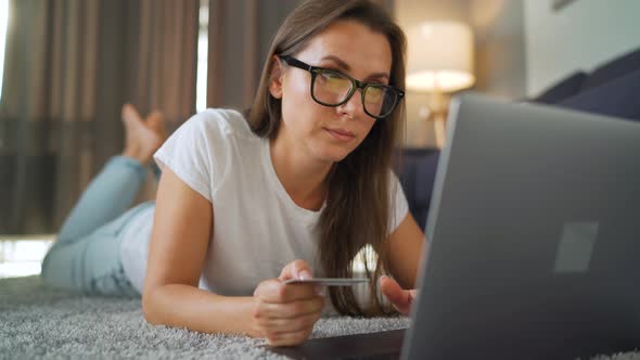 Woman with Glasses Is Lying on the Floor and Makes an Online Purchase Using a Credit Card and Laptop