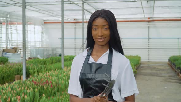 Beautiful Young African Girl in a White Shirt and a Black Leather Apron Stands with Gardening Tools