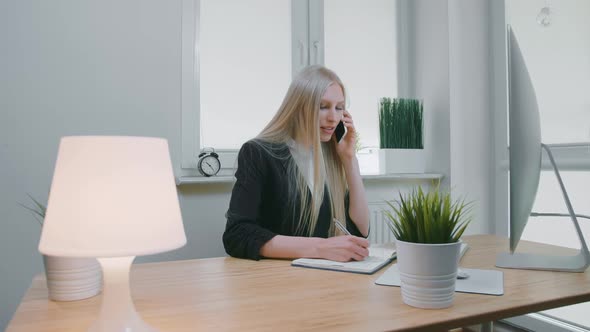 Business Woman Talking on Smartphone in Office. Elegant Young Blond Female in Office Suit Sitting at