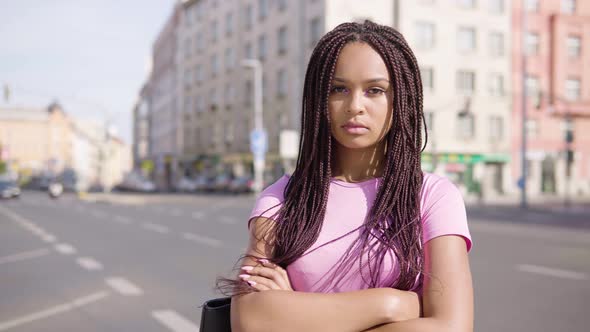 A Young Black Woman Looks Seriously at the Camera in the Street in an Urban Area - a Busy Road