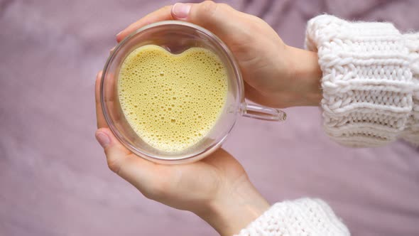 Woman Hands Holding Golden Turmeric Almond Latte In Glass Cup With Heart Shape.