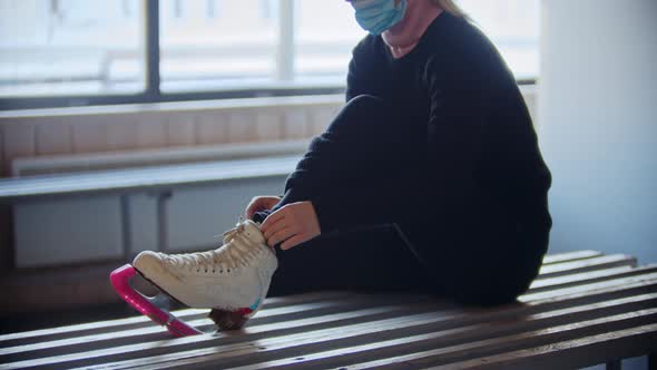 Ice Skating  Young Woman in Medicine Mask Ties Shoelaces on the Skates in Locker Room