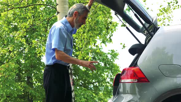 Senior Man Opens Trunk, Rearranges Bags, Then He Goes Away - Trees in the Background