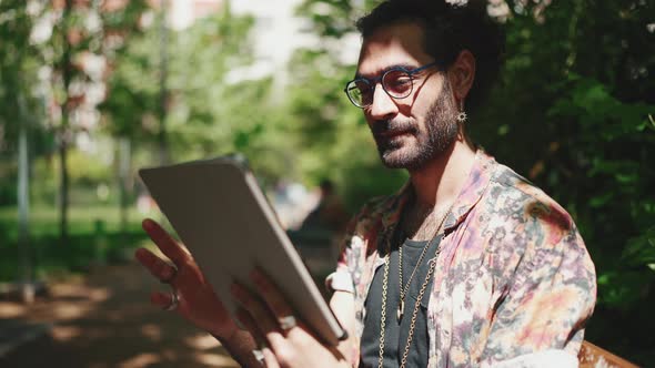 Positive curly-haired bearded man in eyeglasses looking at tablet