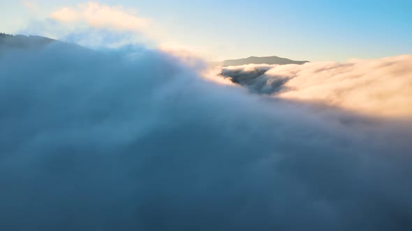 Aerial view of landscape above foggy fast moving clouds covering mountain hills at sunset.