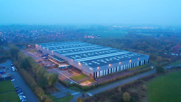 Warehouse at dusk: Aerialing back up, with the lights of the nearby city and roads. Cinematic. Wattr