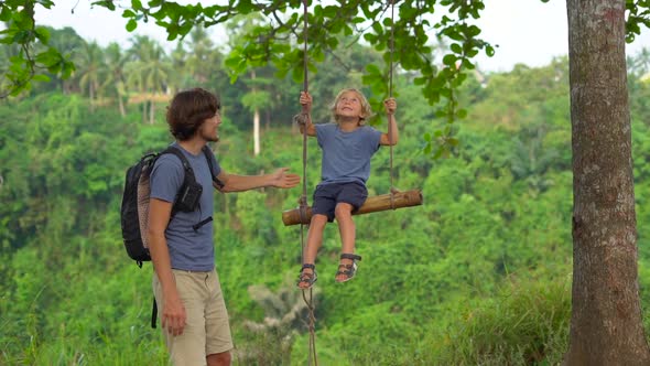 Slowmotion Shot of a Young Man Swinging His Little Son on a Swings in a Tropical Park