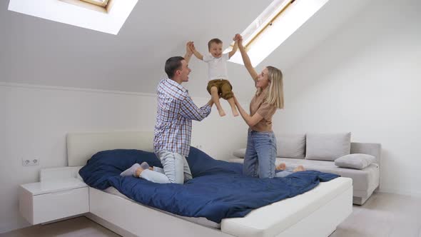 Happy Young Adult Parents with Child Playing Jumping on Bed in Bedroom