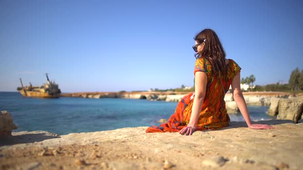 Beautiful Young Woman in a Dress Sitting By Rocky Shore on the Background of Picturesque View