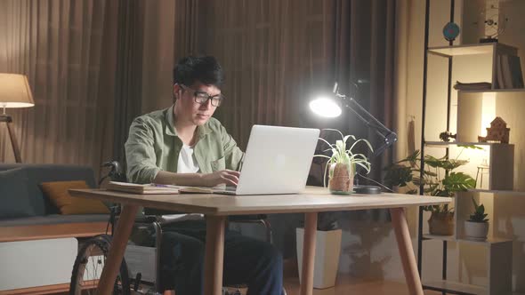 Asian Man Sitting In A Wheelchair While Working On Laptop Computer At Home, Night Time