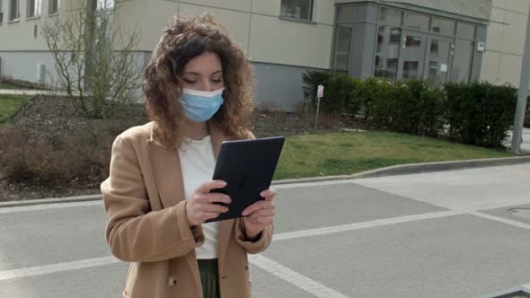 Beautiful Woman in a Protective Mask with a Tablet in Hand Walks Along a Deserted City Street