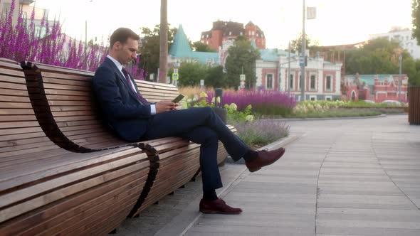 Young Businessman in Official Dark Blue Suit Sits on Bench in Park Before Meeting with Partners and