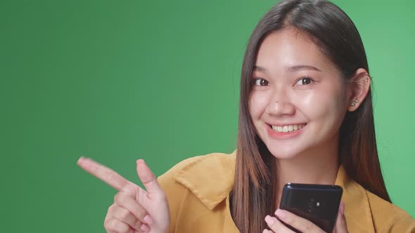 Asian Woman Flipping Through The Phone With A Smile Pointing To The Side In The Green Screen Studio