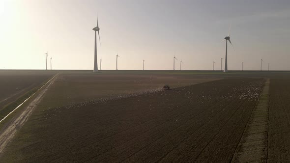 Orbiting around Tractor working on land followed by seagull, Windmill farm Background