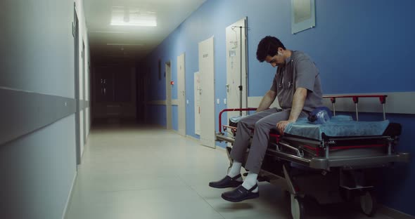 A Doctor Sits on a Medical Gurney and Rubs His Head in Empty Hospital Corridor