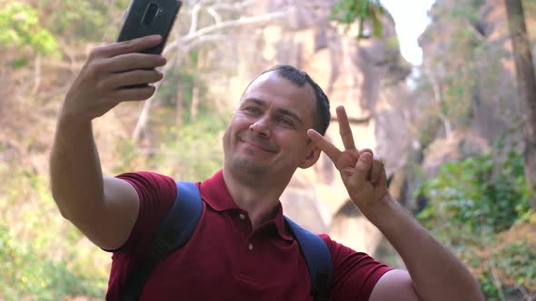 A Smiling Male Traveler Takes Selfies on His Phone Against a Canyon in a Forest Park