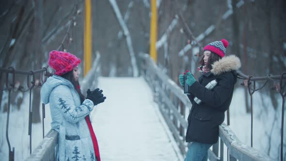 Two Young Women Standing and Having a Talk on the Snowy Bridge - Drinking Hot Drinks 