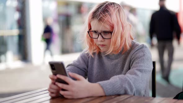 A Caucasian Teenage Girl with Red Hair and Glasses Using a Mobile Phone is Sitting at a Table in a