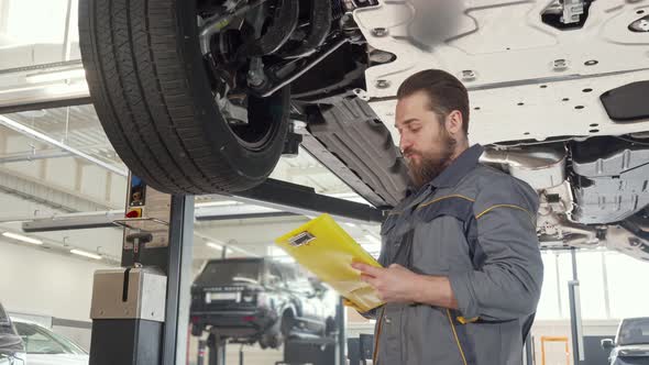 Low Angle Shot of a Bearded Car Mechanic Taking Notes, Examining Lifted Auto