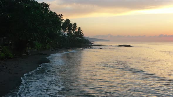 sun rising over horizon at dawn with calm ocean waves crashing on tropical bali beach, aerial