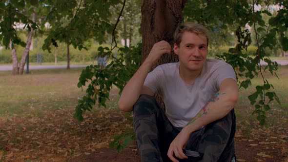 Close Up of Young Man Rests Sitting Near Tree in Parkland