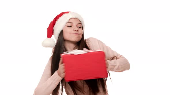 Portrait of Pleased Teen Girl Having Good Mood Dancing in Santa Claus Red Hat Holding Christmas Gift