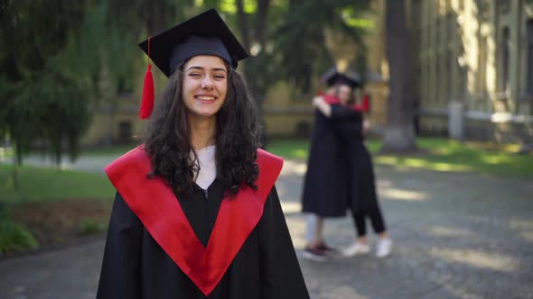 Portrait of Joyful Caucasian Graduate Student Looking at Camera Smiling with Blurred Couple Hugging