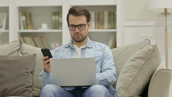 Young Man with Smartphone Working on Laptop at Home