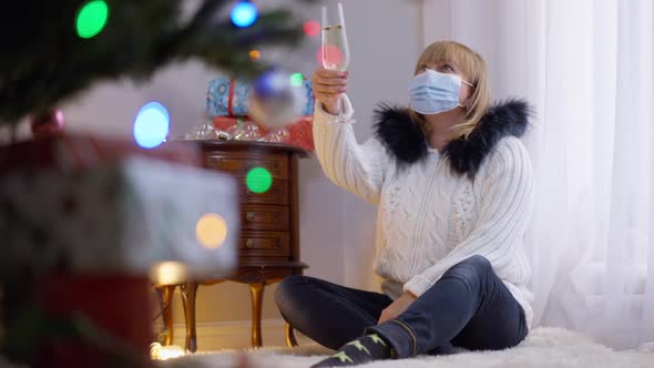 Wide Shot Lonely Woman in Covid19 Face Mask Toasting Champagne with Decorated Christmas Tree Sitting