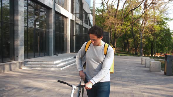 Courier with Yellow Bagpack Walking with Bicycle in City Park and Using Smartphone Slow Motion